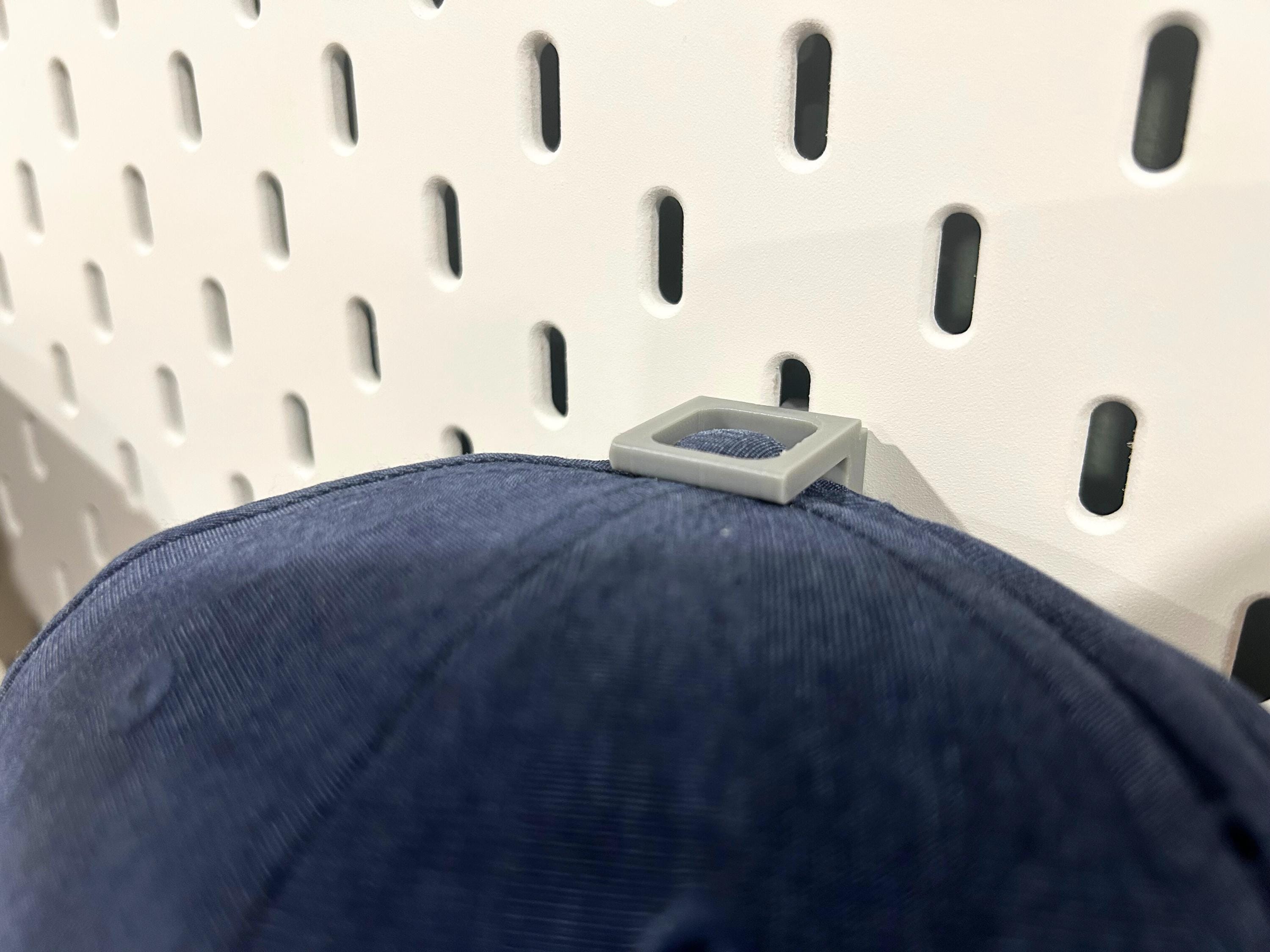 A blue baseball cap with a plastic hook attached to the top of the cap to hold it on a pegboard.